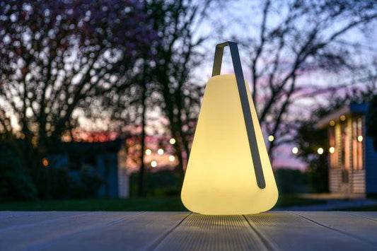 A glowing, cone-shaped, weatherproof lantern with a handle graces the patio at dusk. String lights and silhouettes of trees and a house outline the background against a purple and pink sky. The Brisks Extreme Lounging B-Bulb Outdoor Light ensures your evenings are both magical and practical.