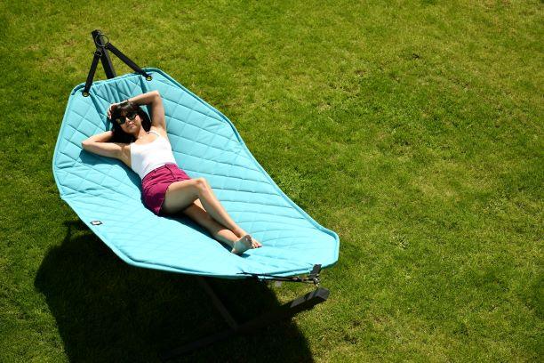 A person is lounging on a large, blue Extreme Lounging B-Hammock by Brisks placed on green grass. They are wearing a white sleeveless top, pink shorts, and sunglasses, with one arm behind their head and the other resting on their stomach, relaxing under the sun in a durable and comfortable setup.