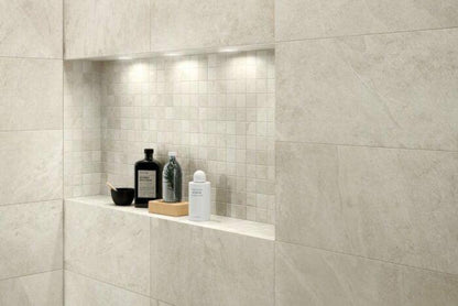 A recessed shower shelf with various toiletry items is set against a light beige tiled wall. The shelf, accented with Brisks Frame Glen Porcelain Paving Slabs and small square tiles, is illuminated by three overhead lights. Items on the shelf include bottles, a soap dish, and a small round container made from sustainable sourcing.