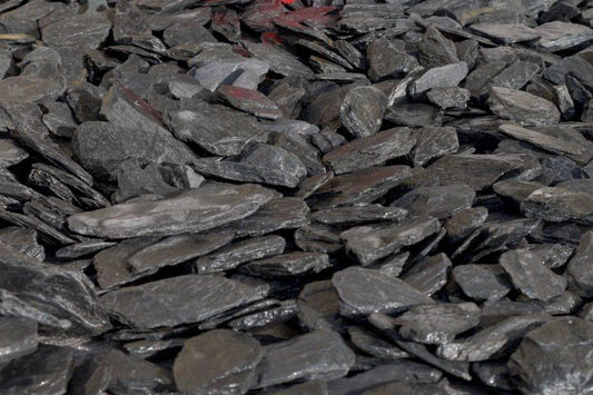 A close-up image of a large pile of 40mm Graphite Grey Slate Chippings by Brisks. The stones are irregularly shaped, flat, and layered on top of each other, creating a rough and textured surface. Perfect for landscaping projects, the image highlights the dark, shiny, and slightly reflective quality of the slate.