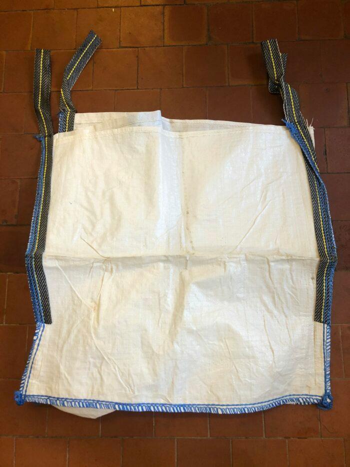 A large, white, durable woven polypropylene bag from Brisks lies flat on a brick-tiled floor. The Empty Bulk Bag has four blue-striped, reinforced lifting loops at each corner and blue stitching along its seams. This sustainable reusable bag appears to be empty and unused.