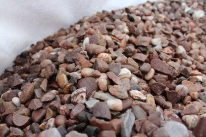 A close-up view of a pile of small, mixed-color gravel stones, including 20mm Staffordshire Pink Gravel by Brisks, with hues of brown, gray, white, and red. The decorative garden gravels are irregular in shape and size, creating a textured surface. The background is a blurred white surface. Perfect for landscaping projects.