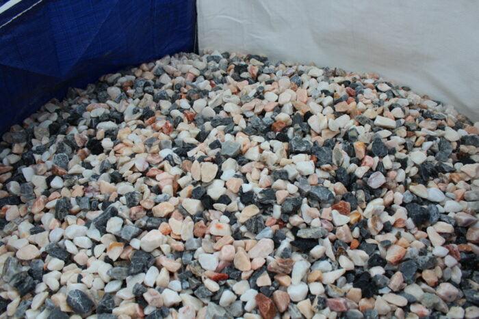A large container filled with small, tumbled stones in various colors, including white, black, pink, and gray. The stones are irregularly shaped and appear smooth with a slightly polished surface, making them perfect for garden landscaping. Among them are some beautiful 20mm Polar Pink Marble Chippings from Brisks.