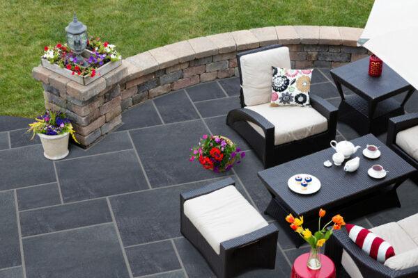 An outdoor patio with Brisks Black Limestone Paving Slabs features cushioned wicker furniture, a sleek black coffee table with a tea set, and colorful potted flowers. A curved brick wall adorned with more flowers borders this elegant modern space, while a green lawn extends beyond the patio.