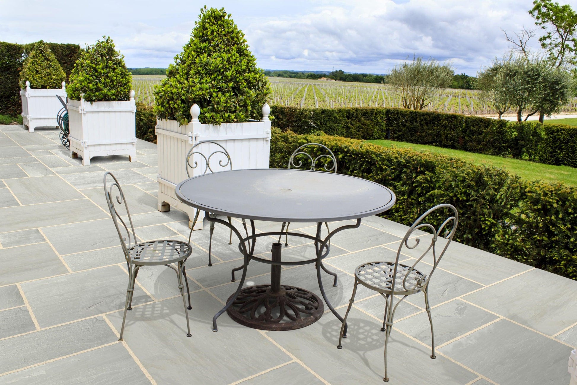 An outdoor patio with a round metal table and three matching chairs sits on a floor paved with Brisks Kandla Grey Sandstone Paving Slabs. In the background, manicured hedges, large white planters with green shrubbery, and a scenic view of fields and trees under a cloudy sky complete the serene setting.