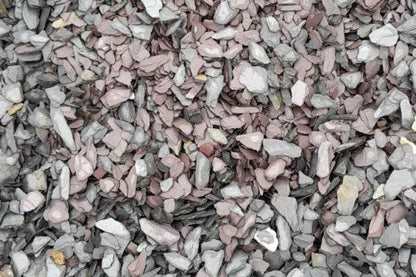 A close-up view of a pile of grey and pinkish stones and pebbles, resembling 20mm Mixed Blue & Plum Slate Chippings from Brisks, in various sizes and shapes. The Blue & Plum Slate hues create a textured and rugged surface, enhancing the raw, natural aesthetic perfect for landscaping projects.