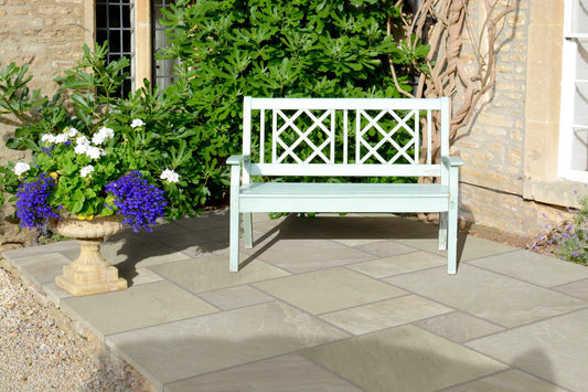 A light green wooden bench is charmingly placed on a stone patio, featuring Brisks Fossil Mint Sandstone Paving Slabs, in front of a stone house with an ivy-covered wall. To the left of the bench, there is a decorative stone planter filled with green foliage and white and purple flowers, enhancing this inviting outdoor space.