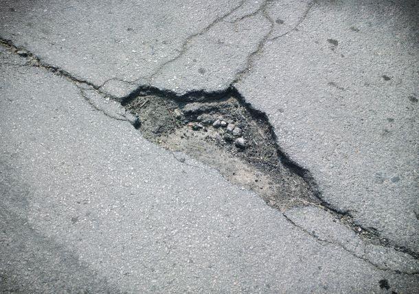 An asphalt road with a large pothole in the center awaits Brisks Cold-Lay Patching Tarmac. The edges of the pothole are cracked, revealing exposed gravel and dirt. The surrounding road surface appears worn and weathered, calling for immediate temporary road repair.