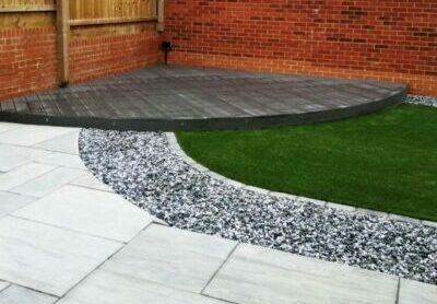 A backyard with a circular wooden deck in the corner, a grassy area separated by a strip of Brisks 20mm Polar Black Ice Chippings, and a light grey stone pathway curving in the foreground. The backdrop consists of a red brick wall and a wooden fence.