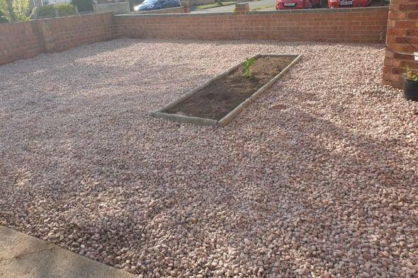 A front yard covered in Brisks 20mm Staffordshire Pink Gravel with a rectangular garden bed in the center containing a small, newly planted tree or shrub. The garden bed is bordered with paving stones. Brick walls and a red car are visible in the background, showcasing careful landscaping.