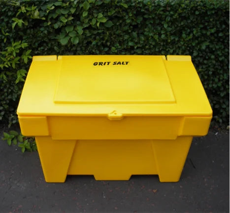 A large, 200-litre Grit Salt Bin by Brisks made of durable polyethene with a hinged lid sits on the ground in front of a lush, green hedge. The words "GRIT SALT" are printed on the top of the impact-resistant lid in bold black letters.