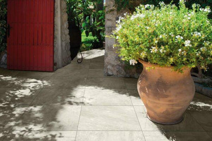 A large terracotta plant pot with lush greenery and white flowers sits on a patio adorned with Brisks Ultra Aspen Bianco Porcelain Paving Tiles in front of a red wooden door and stone wall. Shadows of leaves create patterns on the paving tiles under a sunny, clear sky, perfect for outdoor settings.