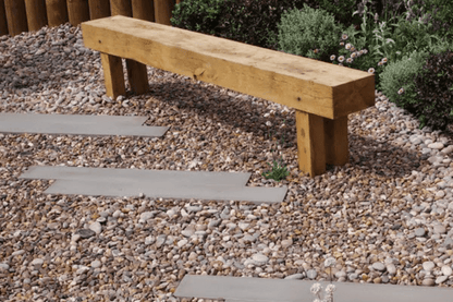 A minimalist wooden bench stands atop a bed of Brisks 10-20mm Shingle Gravel in a garden. The gravel provides excellent drainage, interspersed with rectangular concrete stepping stones leading through the space. Green shrubs and flowering plants are visible in the background, enhancing the landscaping.