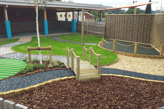 A landscaped garden features a small wooden bridge over a dry gravel stream, winding stone pathways, and an assortment of greenery including a small tree. Brisks Playground Bark lines the pathways, while the space is bordered by a wooden fence with a building and covered area in the background.