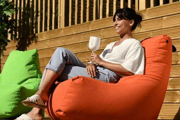 A woman relaxes on an orange Brisks Extreme Lounging B-Bag Mighty outdoors, smiling and holding a wine glass. She is dressed casually in a white top and light blue pants. In the background, there is a green bean bag chair and a wooden fence.