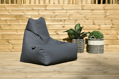 A dark grey Extreme Lounging B-Bag Mighty by Brisks made from premium quality fabric sits on a wooden deck in an outdoor area. Behind it, there is a wooden fence and two potted plants, one with green leafy foliage and the other with a neatly trimmed round shrub. This setup promises extreme lounging in comfort and style.
