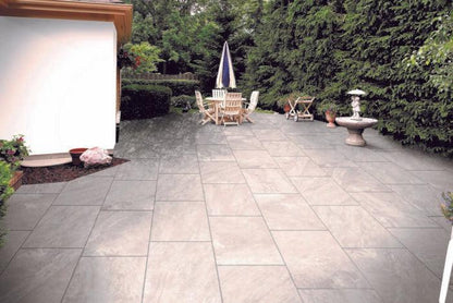 A spacious backyard patio with large, durable Ultra Aspen Grigio Porcelain Paving Tiles by Brisks. Centered is a wooden dining table with chairs around it and a large umbrella. To the right, there's a birdbath, and the area is surrounded by lush green trees and bushes. A white building is adjacent.