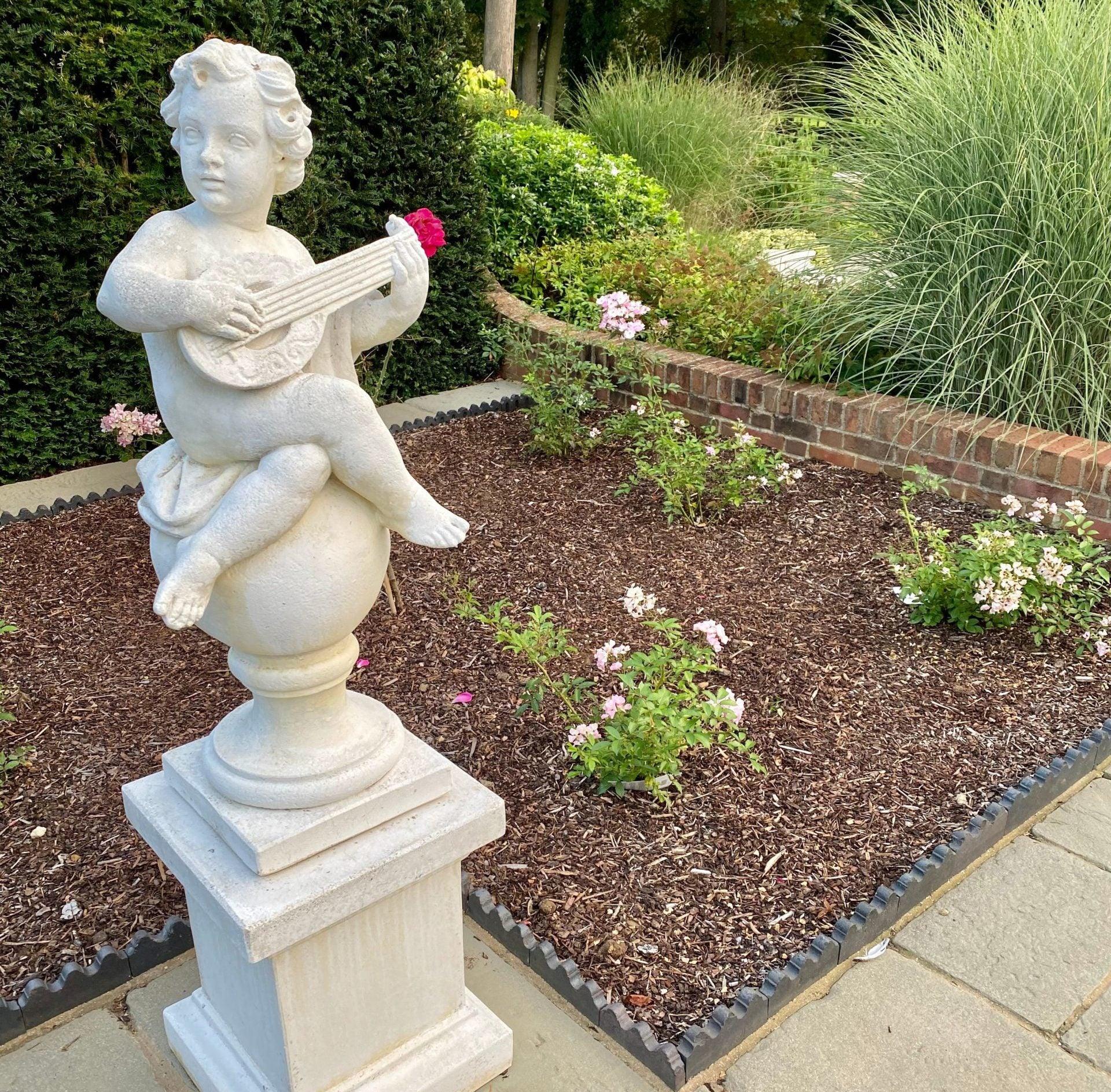 A stone statue of a cherub playing a guitar is placed in a well-maintained garden. The cherub is perched on a sphere atop a pedestal, surrounded by Brisks Bark Mulch for plant protection. Behind the statue, there is a flower bed with green plants, pink flowers, and a brick border.
