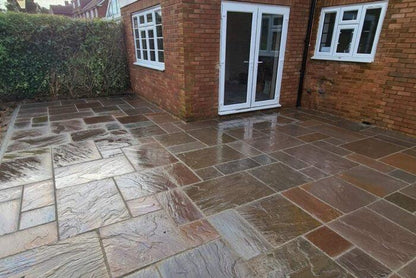 A neatly laid rectangular patio with Brisks Autumn Brown Sandstone Paving Slabs outside a brick house. The patio connects to the house through a set of white double doors and windows above. The area appears wet with a light sheen on the stones. A hedge borders the area on one side, enhancing its natural tones.