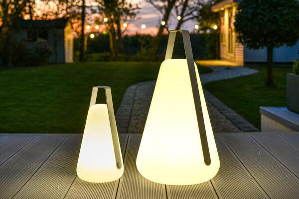 Two cone-shaped, weatherproof outdoor lights with glowing LEDs sit on a wooden deck during twilight. Both Brisks Extreme Lounging B-Bulb Plus Outdoor Lights have handles on top and are positioned outdoors with trees and a building in the background, illuminated softly by nearby lights.