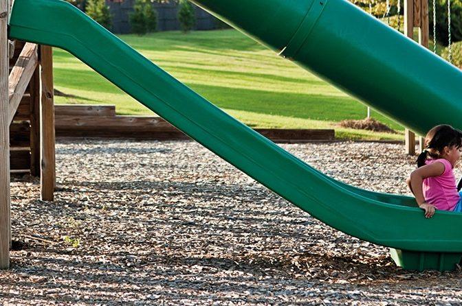 A young girl wearing a pink top slides down a green playground slide on an outdoor play area with grassy surroundings. The ground is covered with Brisks Playground Bark, and the scene includes some wooden structures along with various other playground equipment.