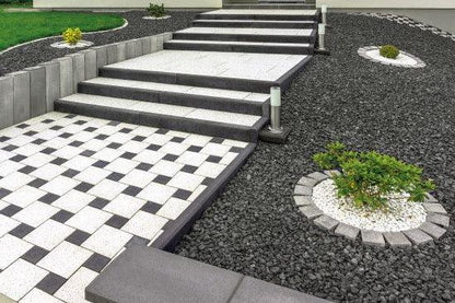 A modern outdoor staircase with white and dark grey steps leads up to a higher level. The adjacent pathway showcases a checkered black and white tile design. The landscaped areas feature Brisks 10-20mm Black Basalt Chippings with circular patches of plants surrounded by decorative white gravel.
