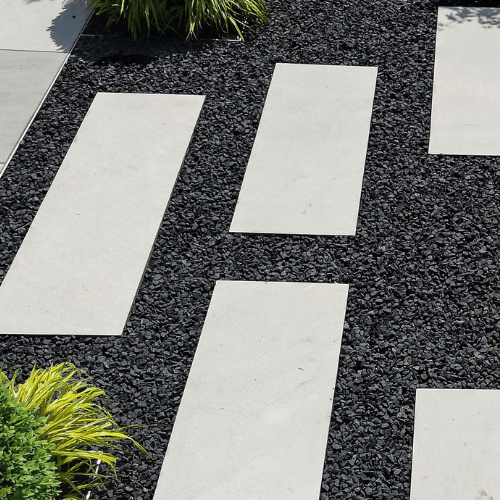 Rectangular white stone pavers are evenly spaced and embedded in a pathway of 10-20mm Black Basalt Chippings by Brisks. The edges of the pathway are bordered with green, leafy plants. The sunlight casts soft shadows, creating a clean and modern outdoor design.
