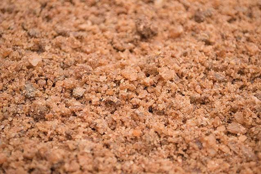Close-up of reddish-brown coarse particles resembling Brisks Brown Rock Salt. The texture appears rough and crumbly, with a mixture of small and slightly larger granules. There are no distinguishing features or items within the image besides the granular material, suitable for commercial and residential use in thawing ice and snow.