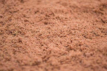 A close-up view of red-brown granular material spread out evenly, resembling Brisks 0-2mm Red Building Sand or fine gravel. The texture appears consistent throughout the image with small, uniform particles.