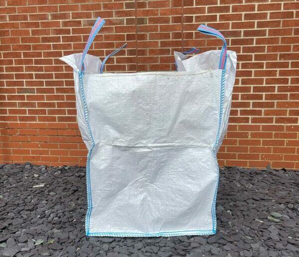 A large, durable woven polypropylene bag with blue stitching stands open and empty on a surface of dark slate chips. The Brisks Empty Bulk Bag, featuring four blue lifting loops at the top, is set against a red brick wall background.