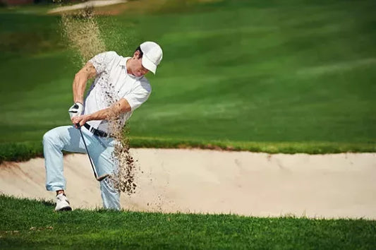 A golfer in a white shirt and white cap is hitting a shot from the sand bunker on a lush green golf course. The Brisks Golf Course Bunker Sand, known for its non-staining property, visibly flies up into the air from the force of the swing.