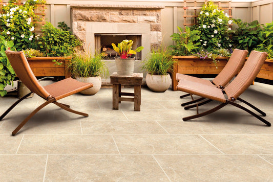 Outdoor patio seating area with Brisks Carmen Beige Porcelain Paving Slabs, wooden lounge chairs with brown cushions, and a small wooden table. A potted plant with colorful leaves sits on the table. The background features stone and wooden planters with lush green plants, showcasing well-designed outdoor spaces.