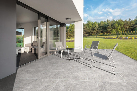 A spacious outdoor patio features several modern chairs and a glass table on Brisks Carmen Grey Porcelain Paving Slabs. The slip-resistant, grey tiled floor adds a touch of elegance. The sliding glass door opens into a contemporary living room. Beyond the patio, a well-manicured lawn extends towards lush trees under a blue, cloudy sky.
