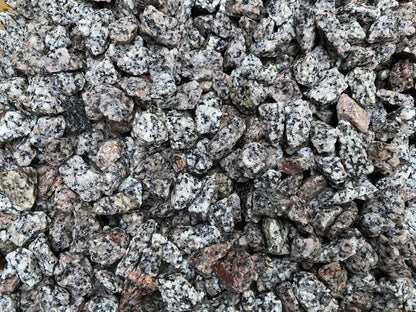 A close-up image of a large pile of coarse, rough Brisks 20mm Black and White Granite Chippings. The stones are irregularly shaped and mostly gray with specks of black and white, showcasing their textured surfaces. These Brisks decorative garden chippings add a rugged elegance to any landscape.