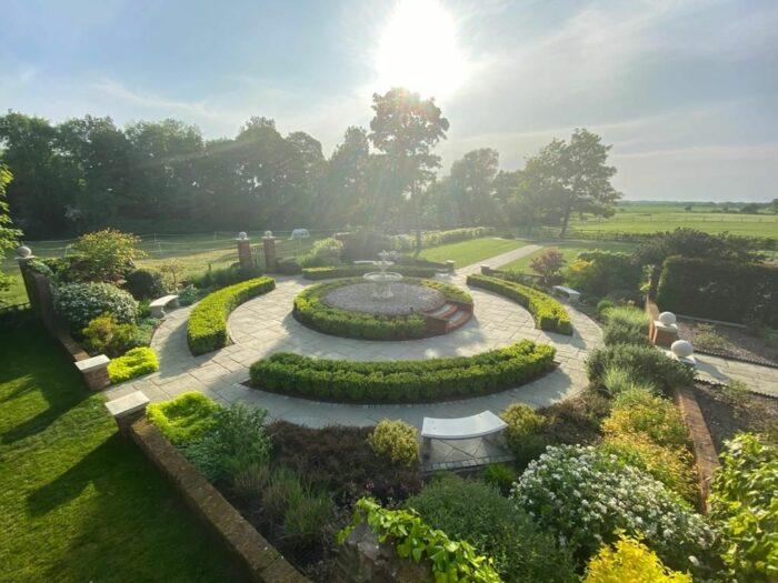 A beautifully landscaped garden with concentric circular paths of Brisks Kandla Grey Sandstone Paving Slabs and lush greenery. In the center is a fountain surrounded by neatly trimmed bushes. Benches are placed along the paths of Brisks Kandla Grey Sandstone Paving Slabs. Tall trees and a field are visible in the background under a bright sun.