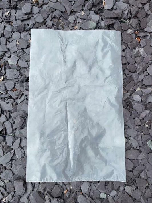 A clear, crumpled plastic sheet lies on a bed of dark grey slate stones. The durable 20kg Empty Rubble Bags from Brisks have visible creases and appear to reflect some light. The slate stones are irregularly shaped and vary slightly in size.
