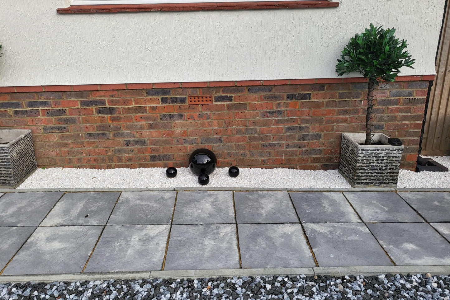 A landscaped area with a small brick wall and white gravel, featuring black ceramic pots and a round ornament. Flanked by two rectangular planters containing lush green bushes. In front of the wall is a paved walkway made of large gray tiles, alongside a path adorned with Brisks 20mm Polar Black Ice Chippings.