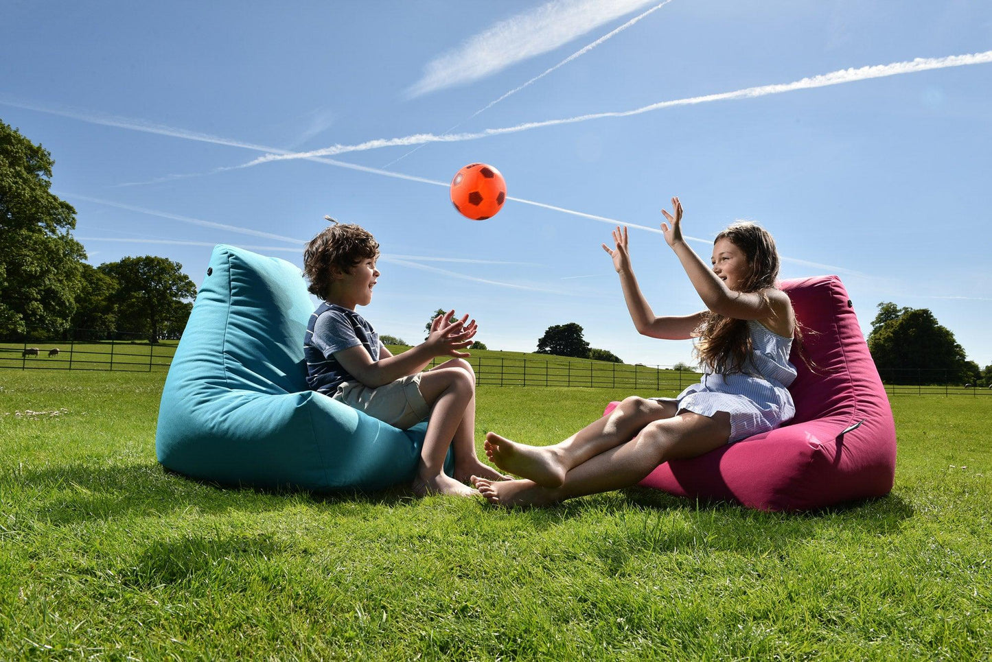 Two children are playing with an orange ball on a sunny day. They are sitting on colorful Brisks Extreme Lounging Outdoor Mini B-Bags on a grassy field. The boy is on a blue one and the girl is on a pink one. Trees and a clear blue sky with contrails can be seen in the background, ensuring durable and comfortable fun.
