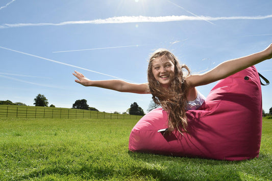 A young girl with long hair joyfully spreads her arms, mimicking flight, as she lies back on a durable and comfortable bright pink Brisks Extreme Lounging Outdoor Mini B-Bag on a lush green lawn under a clear blue sky with a few contrails. Trees and a fence are visible in the background.