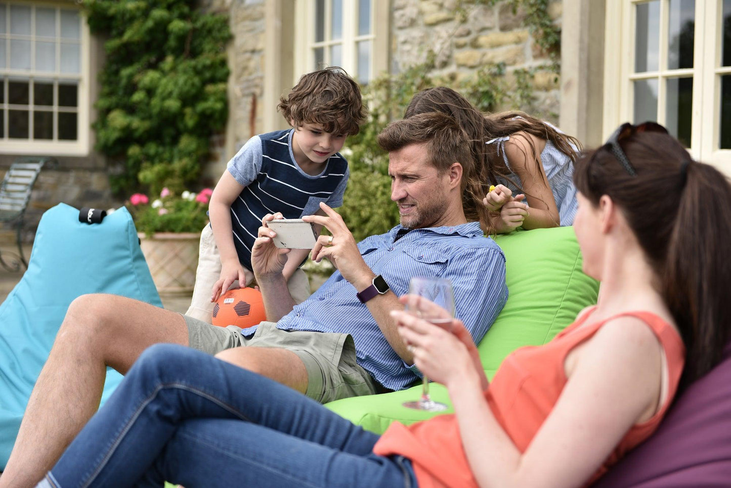A man and a woman lounge on colorful Brisks Extreme Lounging B-Bag Mighty outside a rustic house. The man shows something on his smartphone to two children, while the woman holds a glass of wine and looks on. One child holds an orange ball, and the other stands beside the man.