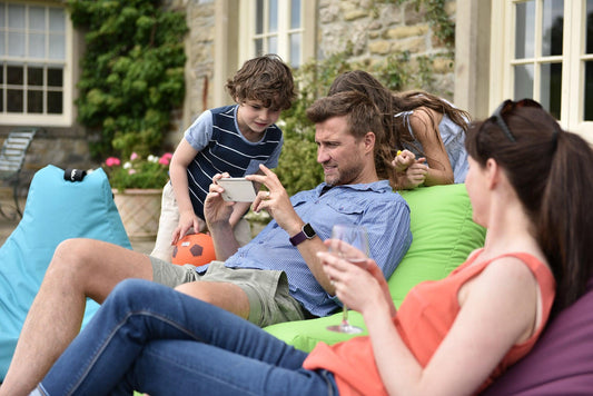 A man and a woman lounge on colorful Brisks Extreme Lounging B-Bag Mighty outside a rustic house. The man shows something on his smartphone to two children, while the woman holds a glass of wine and looks on. One child holds an orange ball, and the other stands beside the man.