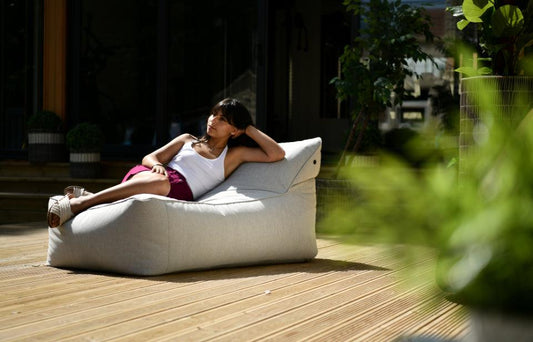 A person is lounging on a large, white Brisks Extreme Lounging B-Bed Pastel, situated on a wooden deck. Dressed in a white tank top and pink shorts with a relaxed expression, they are surrounded by greenery. The image depicts a peaceful garden retreat in a sunny outdoor setting.