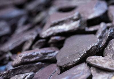 Close-up image of a pile of dark-colored, flat stones with rough edges. The Brisks 20mm Plum Slate Chippings, loosely stacked, show variations in texture and shading with an occasional lilac hue, creating an uneven surface.