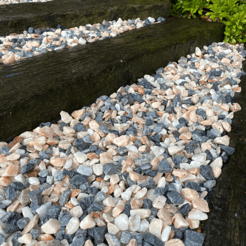 A close-up view of a garden path covered with multicolored gravel stones, featuring a mixture of white, peach, and gray rocks. Wooden steps separate sections of the path, with green shrubs on the side. Brisks 20mm Polar Pink Marble Chippings add an elegant touch to this well-designed garden landscaping.