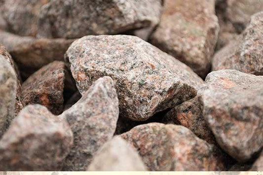 Close-up image of a pile of rough, irregularly shaped gray stones with hints of brown and red tones. The coarse texture and varying sizes indicate this may be 35-50mm Railway Ballast from Brisks, fitting closely together in the frame.