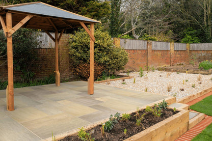 A backyard garden featuring a wooden pergola on a stone patio made of Brisks Raj Green Sandstone Paving Slabs. There are raised garden beds with young plants and a gravel area with sparse vegetation. A brick fence lines the back, with trees and shrubs offering additional greenery, creating an outdoor oasis.