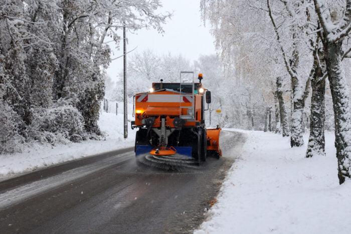A snow plow removes snow from a tree-lined road. The vehicle, adorned in bright orange and blue sections, is actively clearing the slightly snow-covered street using Brisks Brown Rock Salt. Both sides of the road and the trees are heavily covered with snow, indicating a recent snowfall.