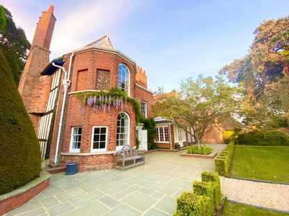 A large, charming brick house with tall chimneys and ivy growing on its facade. The house has several windows and a white-framed door. The front yard, paved with Brisks Kandla Grey Sandstone Paving Slabs, features a bench and is surrounded by well-manicured gardens and trees.