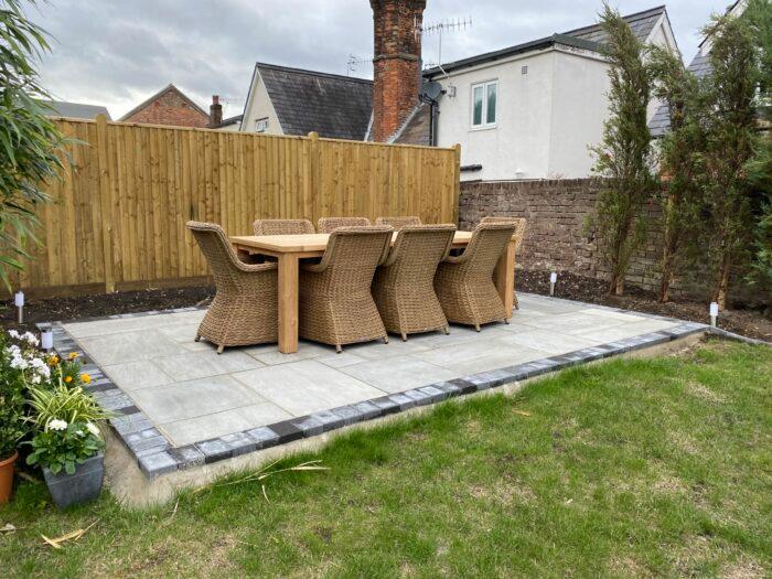 A backyard patio with a wooden dining table surrounded by eight wicker chairs on large Kandla Grey Sandstone Paving Slabs from Brisks. The patio is bordered by a low gray brick edge, with a grassy section in the foreground and a wooden fence along the back, complemented by some plants on the sides.