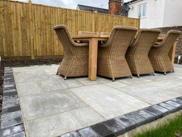 A patio with a set of six wicker chairs and a wooden table is shown. The area is paved with Brisks Kandla Grey Sandstone Paving Slabs, featuring light grey hues and bordered with darker stone bricks. The patio is surrounded by a wooden fence and adjacent to a grassy lawn.